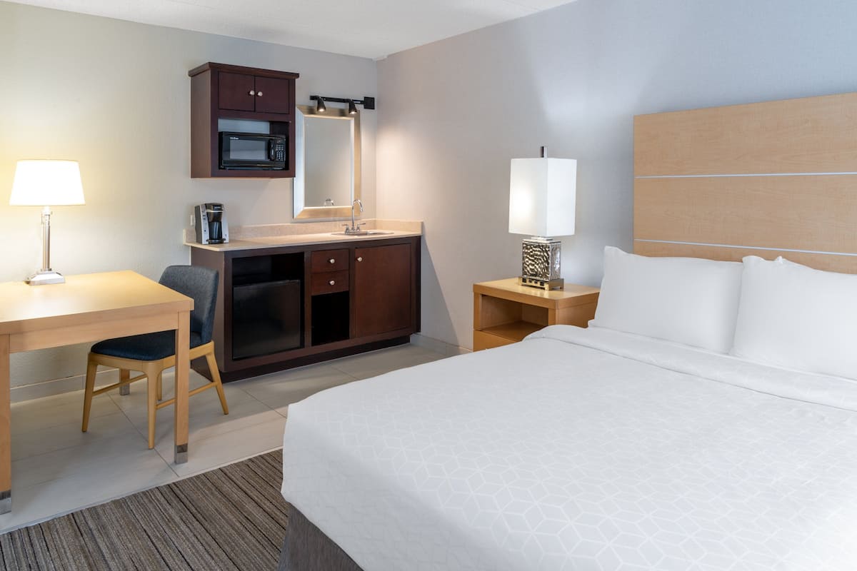 A hotel room with a bed, a desk, a chair, a microwave, a coffee maker, and a nightstand with a lamp.