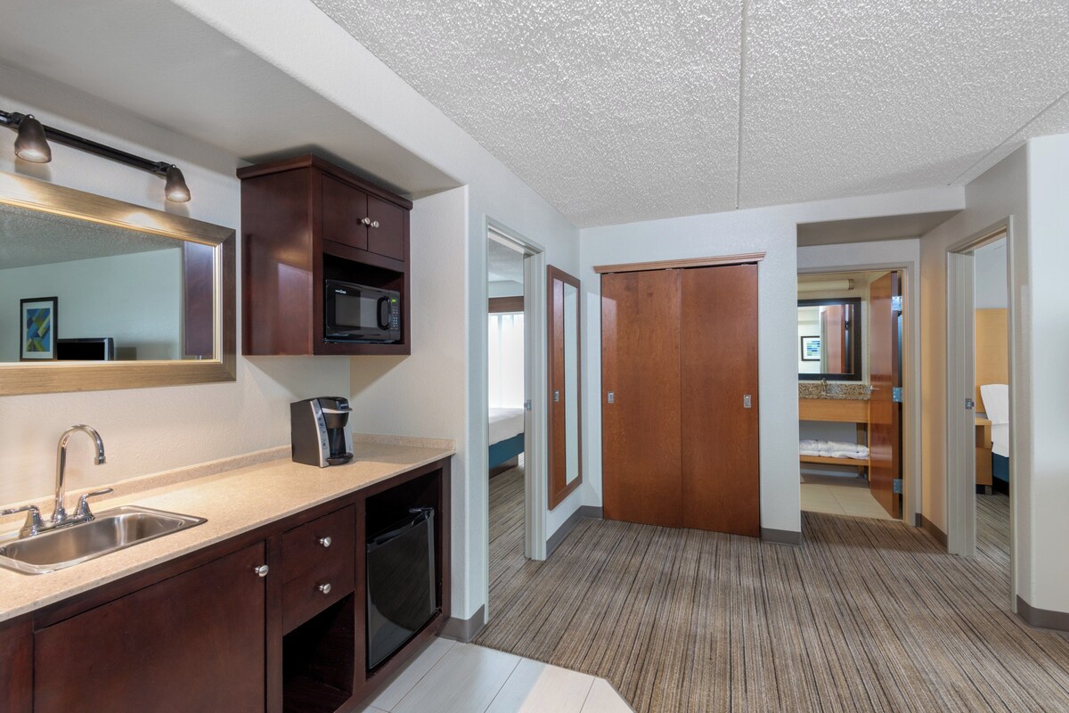 A modern kitchen with a microwave, coffee maker, and sink. There is a wooden cabinet and a mirror above the sink. The floor is carpeted, and there are wooden doors leading to other rooms.