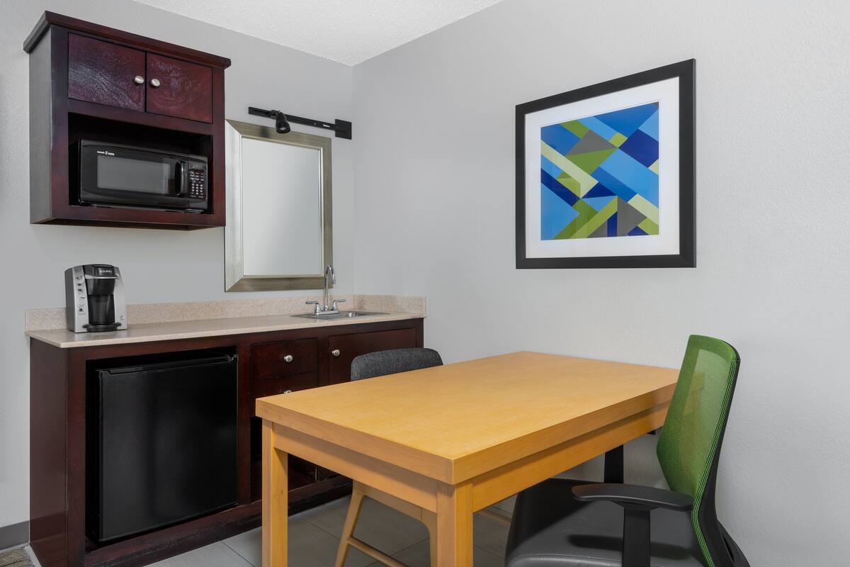 A compact kitchenette with a microwave, coffee maker, and a small refrigerator. There is a wooden table and a green chair. A framed abstract art piece is on the wall.