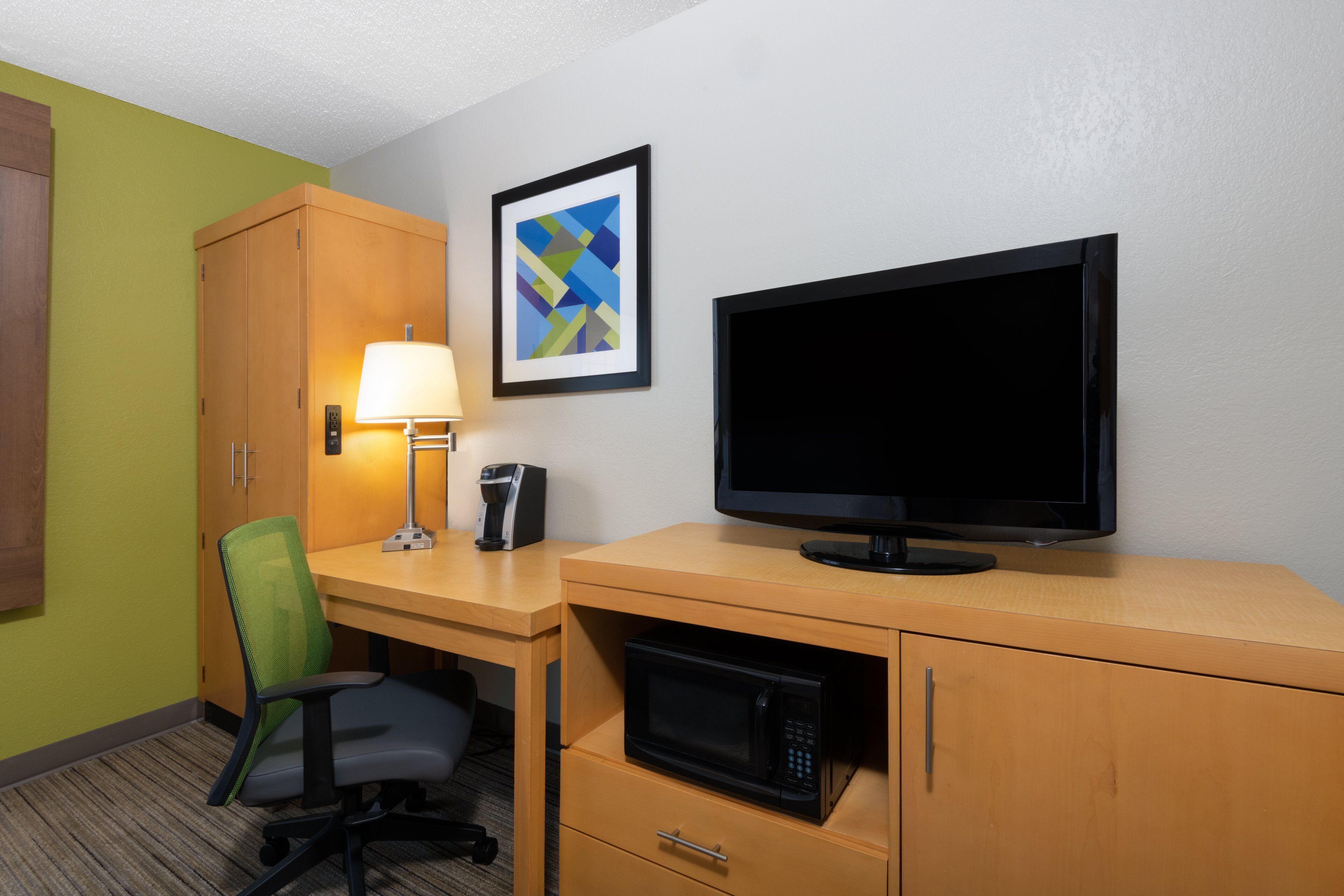 A hotel room with a desk, a chair, a television, a microwave, a lamp, and a framed picture on the wall.