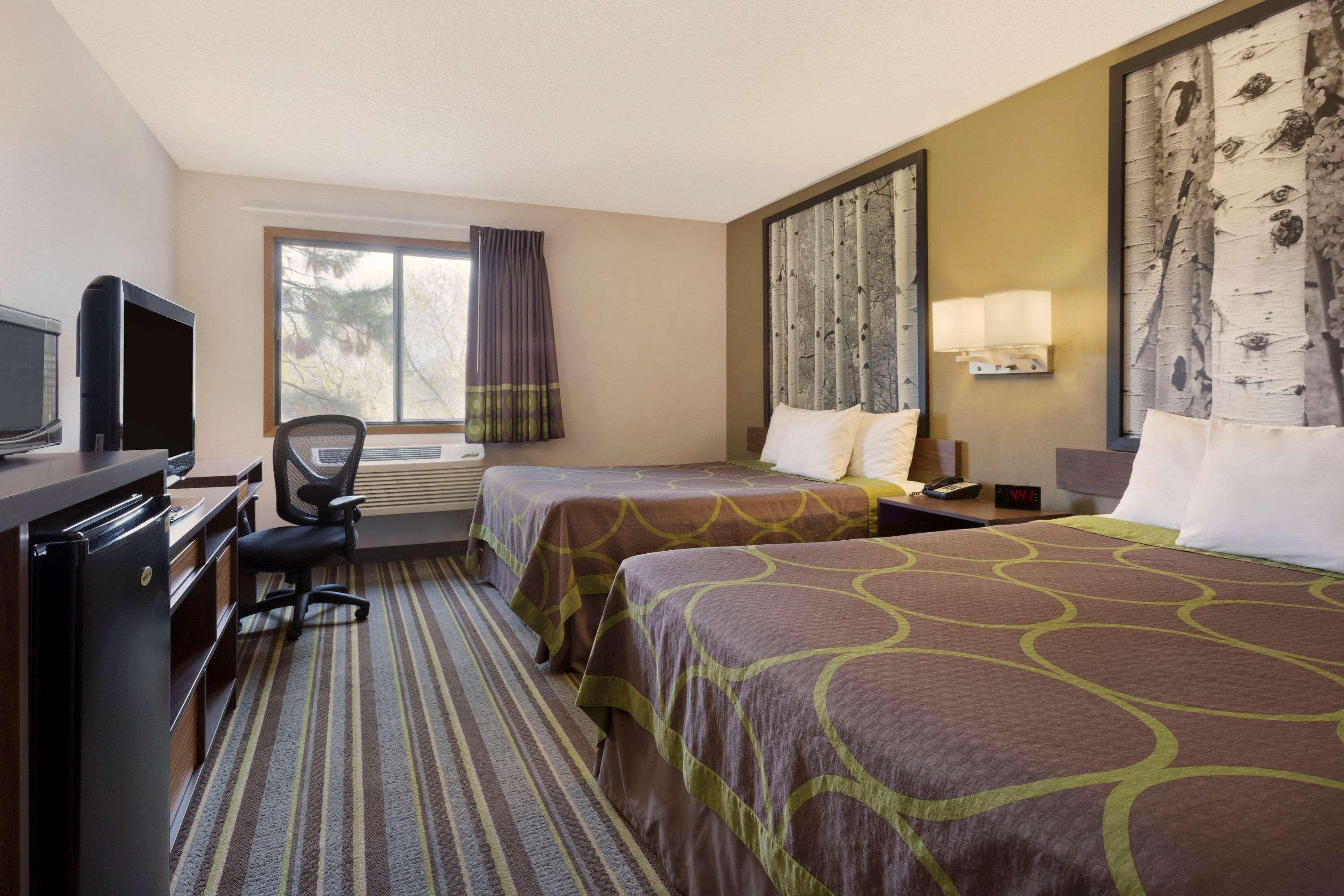 A hotel room with two beds, a desk with a television, a chair, a window with curtains, and a wall-mounted lamp.