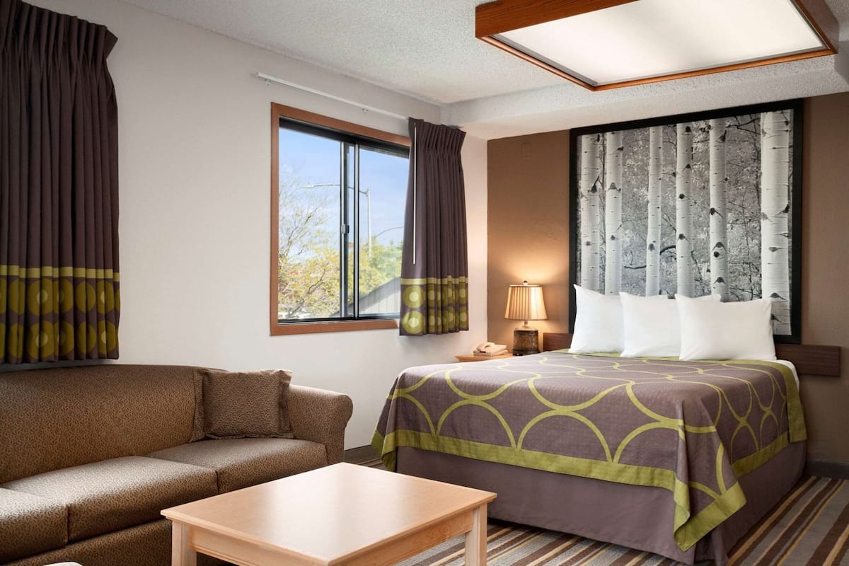 Una habitación de hotel con cama, sofá, mesa, lámpara y ventana con cortinas.