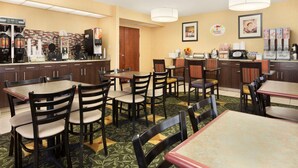 Une salle à manger d’hôtel avec plusieurs tables et chaises, un espace dédié au café avec différents types de machines à café, et un panneau indiquant le buffet du petit-déjeuner sur le mur.