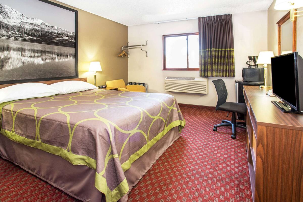 Habitación de hotel con una cama grande, un escritorio con computadora, una silla, un ventanal con cortinas y un aire acondicionado instalado en la pared.