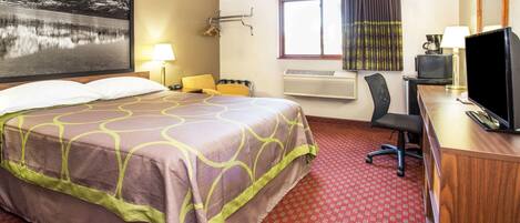 A hotel room with a large bed, a desk with a computer, a chair, a window with curtains, and a wall-mounted air conditioner.