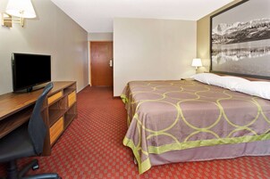 A hotel room with a bed, a desk with a television, a chair, a lamp, and a door.