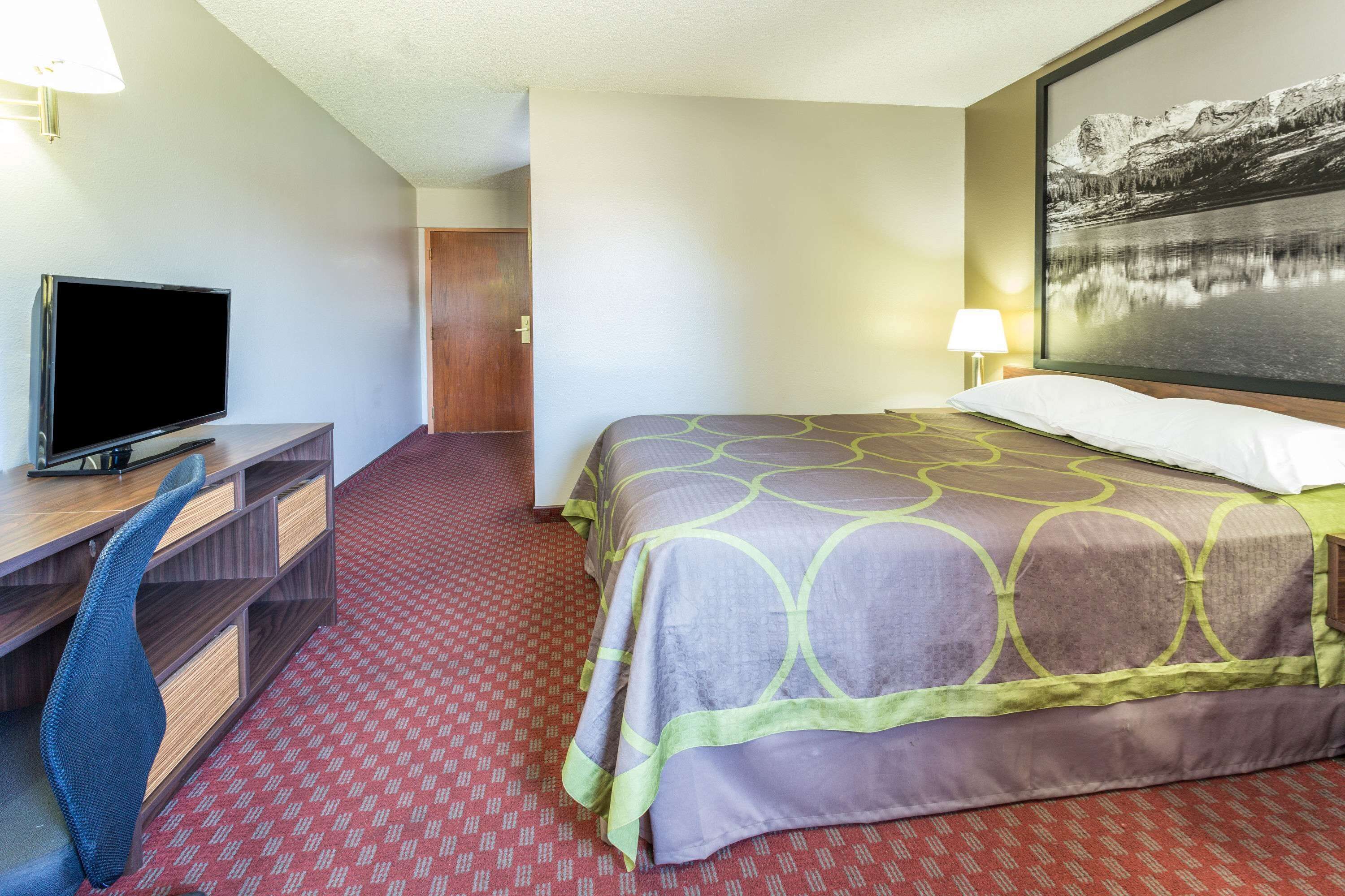 A hotel room with a bed, a desk with a TV, a chair, and a painting on the wall.