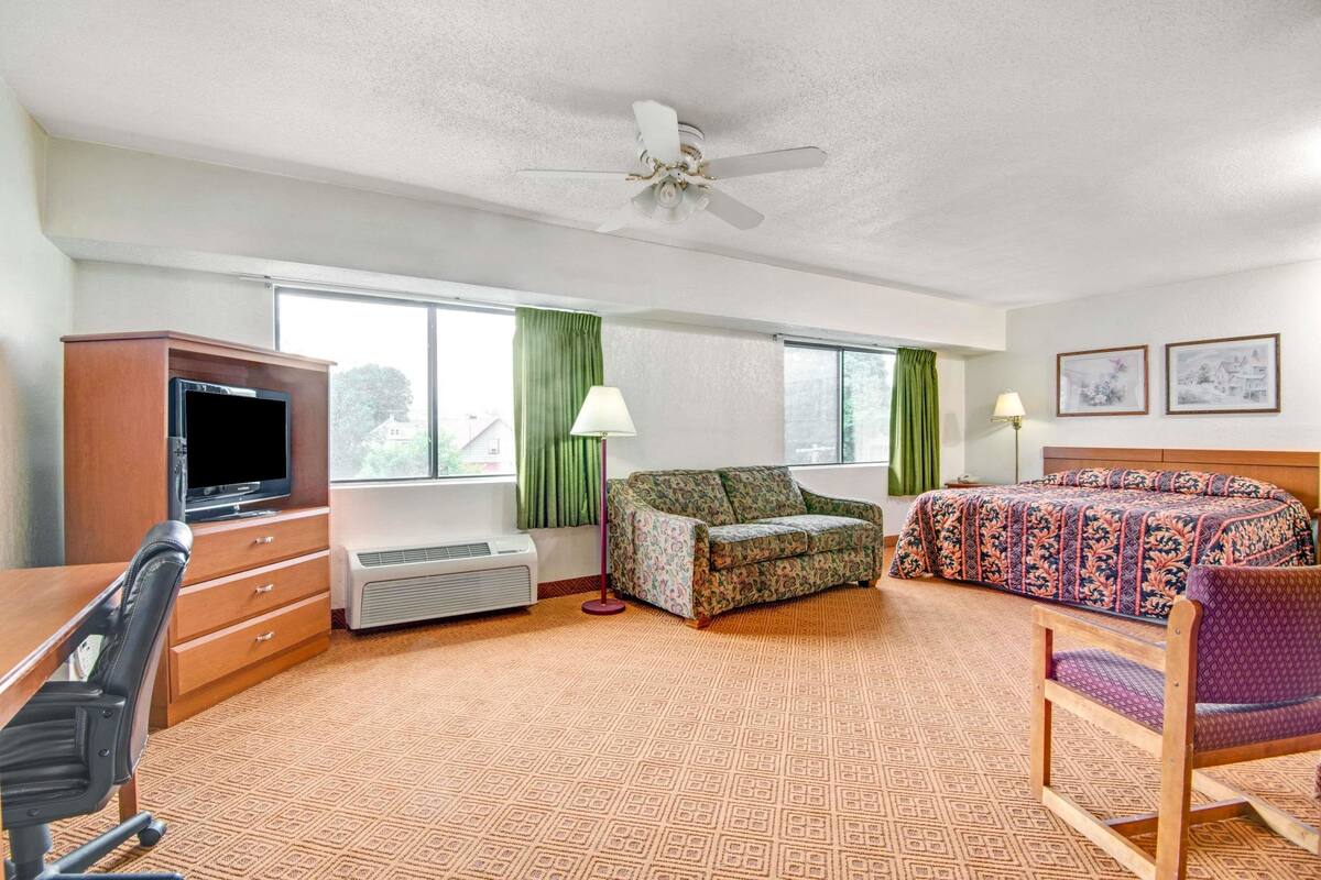 Une chambre d’hôtel équipée d’une télévision à écran plat, d’un canapé, d’un lit avec une couvre-lit à motifs, d’un bureau avec une chaise et d’un ventilateur de plafond.