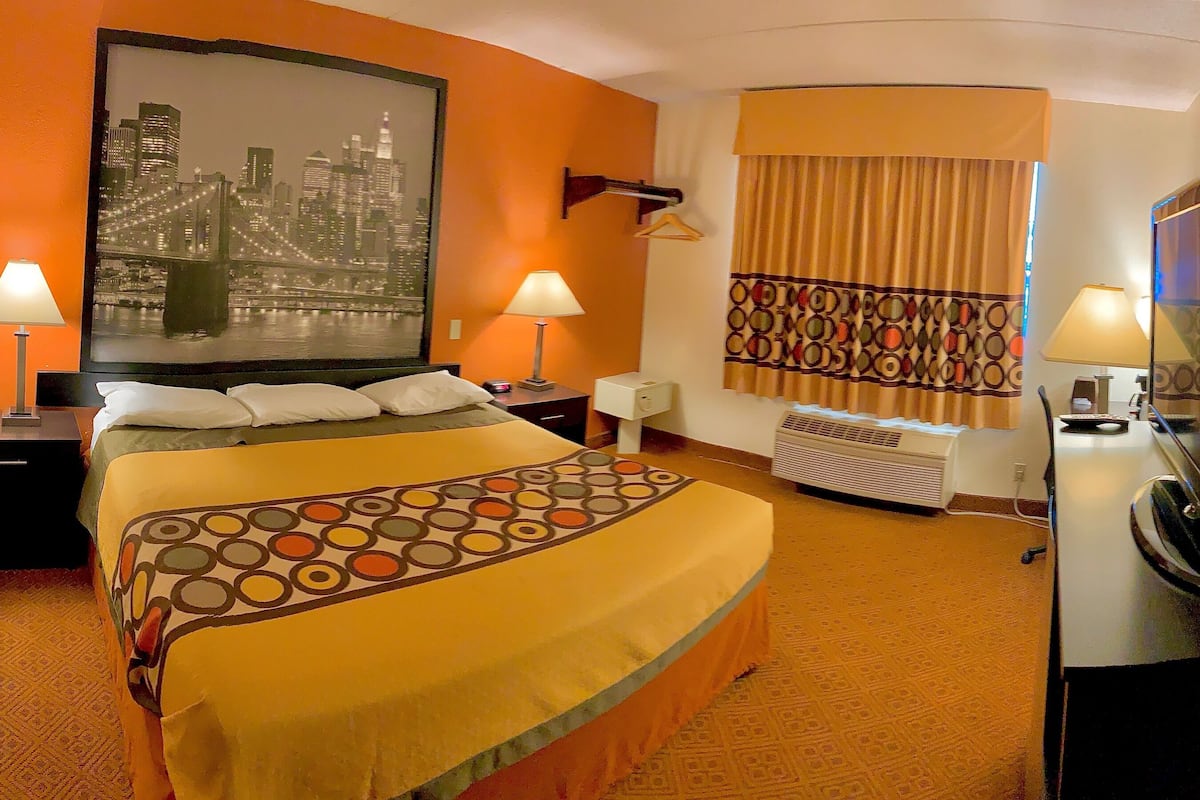 Une chambre d’hôtel avec un lit, deux tables de chevet avec des lampes, un grand tableau représentant un paysage urbain, un rideau à motifs, un appareil de climatisation et un téléviseur posé sur un meuble.