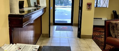 A hotel reception area with a clock, a reception desk, a newspaper rack, and a view of the outside through the door.