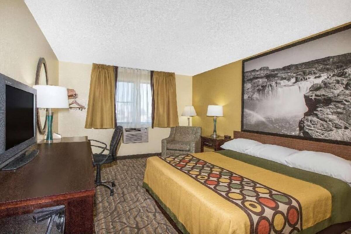 A hotel room with a bed, a desk with a TV, a chair, two lamps, a window with curtains, and a large waterfall painting on the wall.
