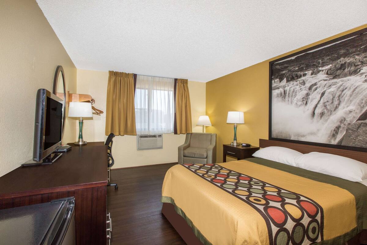 A hotel room with a bed, a desk, a TV, two lamps, a chair, and a large window with curtains.