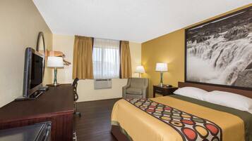 Una habitación de hotel con cama, escritorio, televisión, dos lámparas, una silla y un ventanal con cortinas.