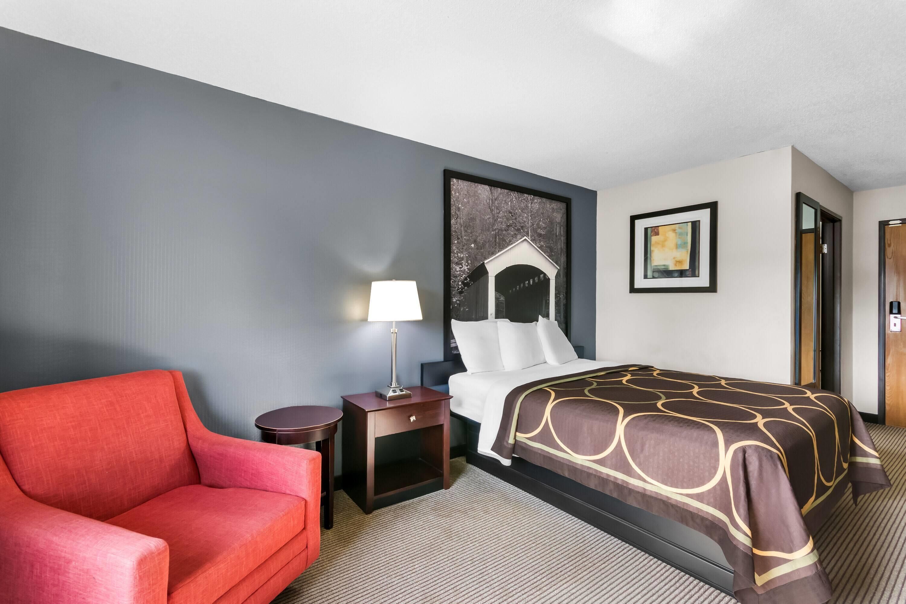 A hotel room with a bed, a red armchair, a small table with a lamp, and a picture on the wall.