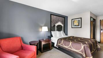 A hotel room with a bed, a red armchair, a small table with a lamp, and a picture on the wall.