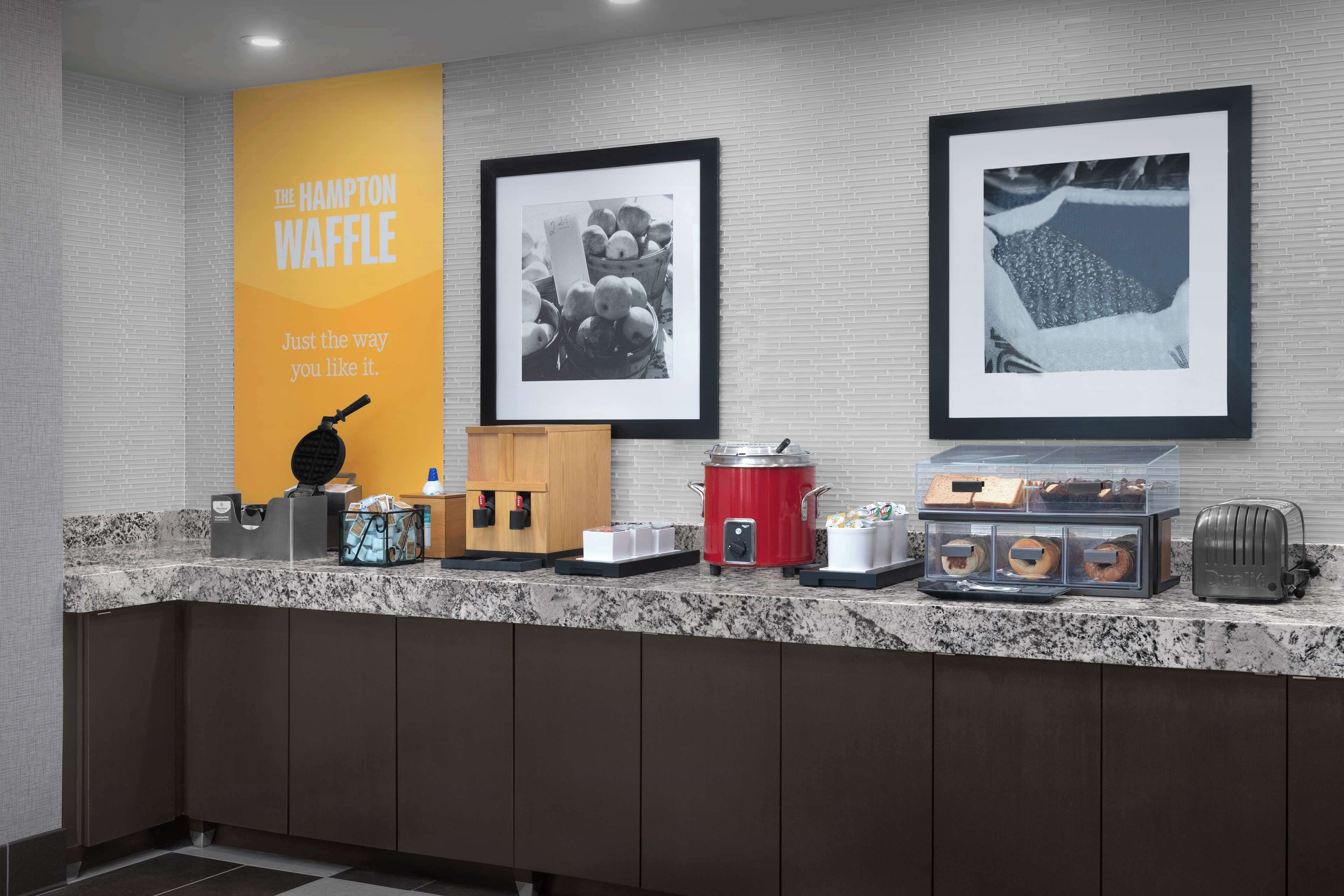 A kitchen counter with a waffle maker, a red slow cooker, a toaster, and a coffee maker. There are framed pictures on the wall above the counter.