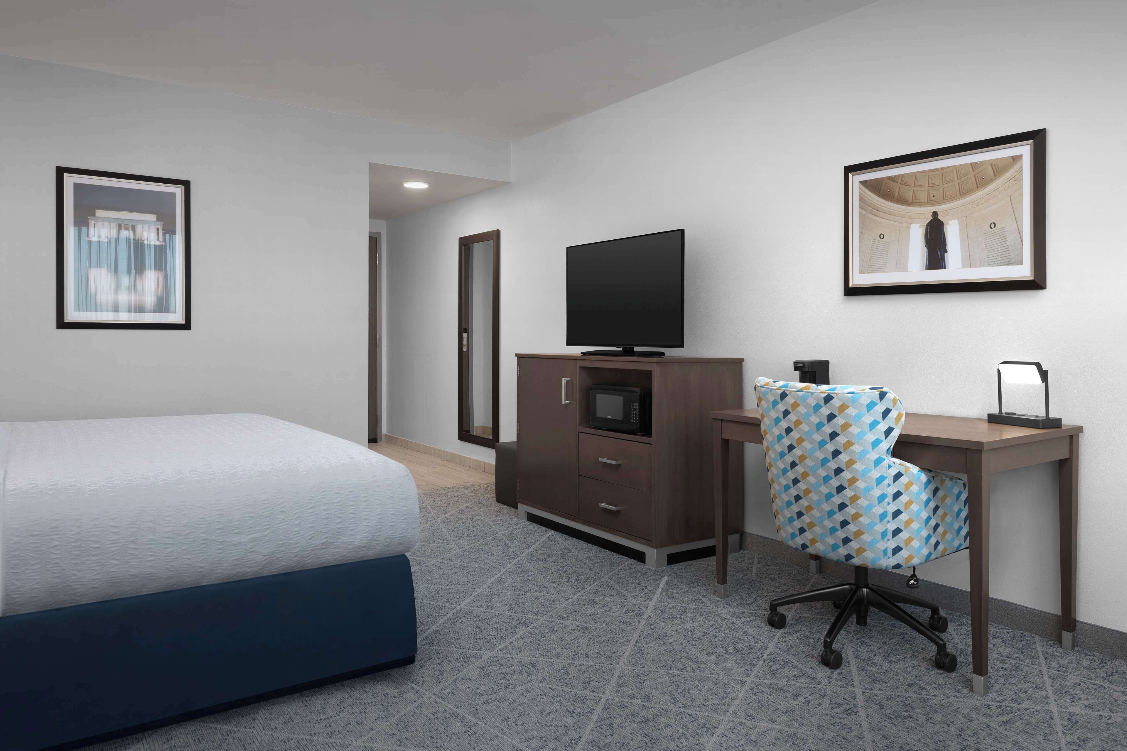 A hotel room with a bed, a desk with a computer, a TV, and a framed picture on the wall.