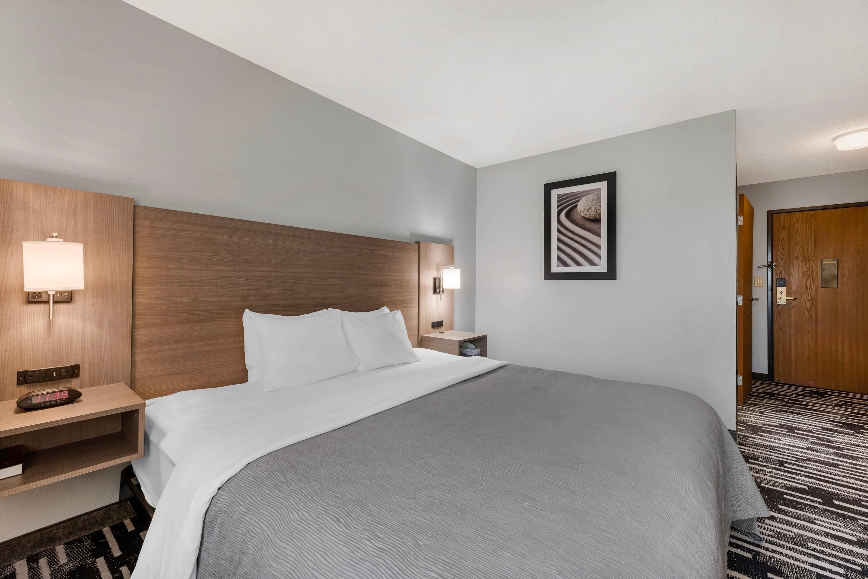 Una camera d'albergo con un letto grande, una testiera in legno, lampade ai comodini, un quadro incorniciato a parete e una porta con maniglia.