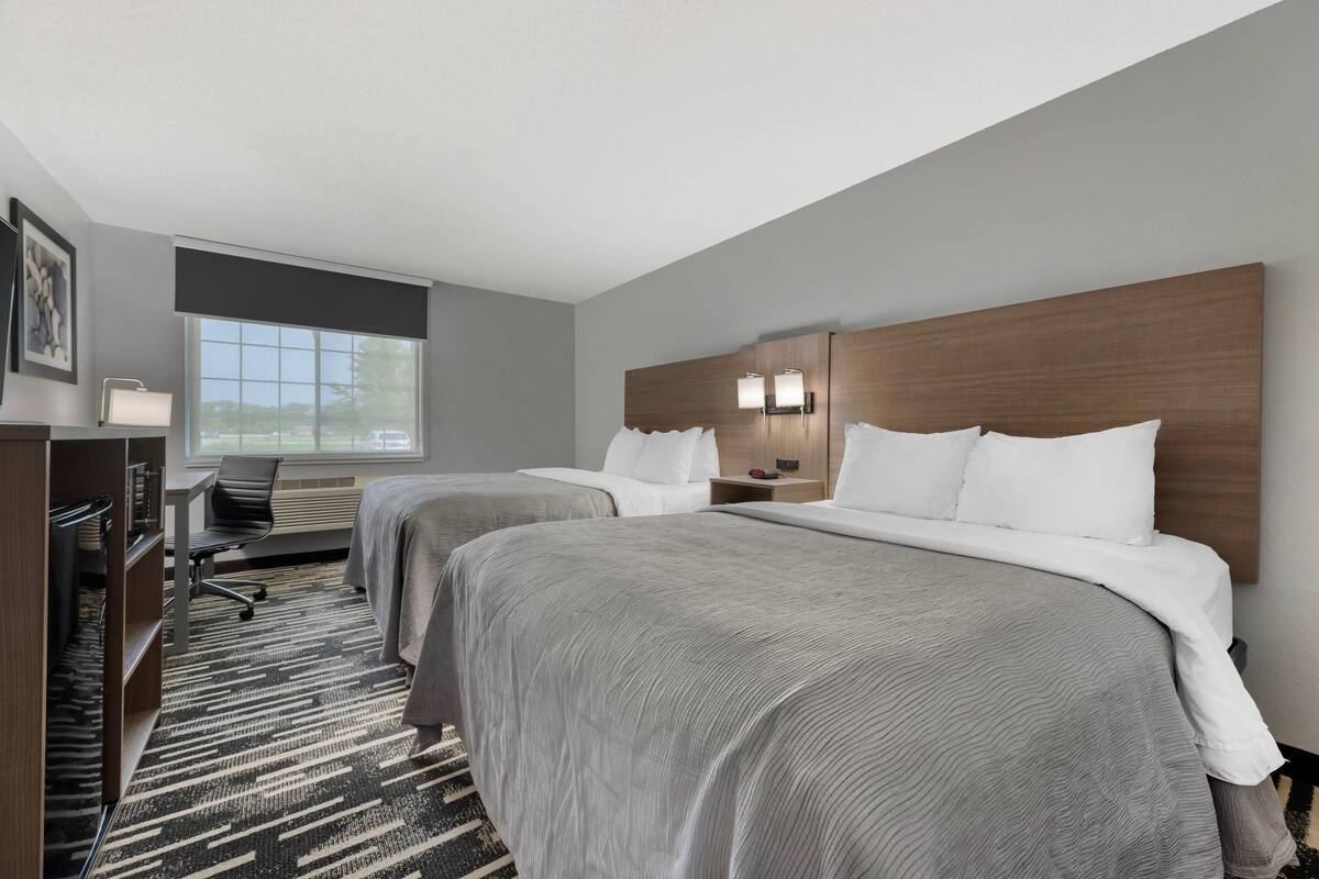 A hotel room with two beds, a desk, a chair, and a large window.