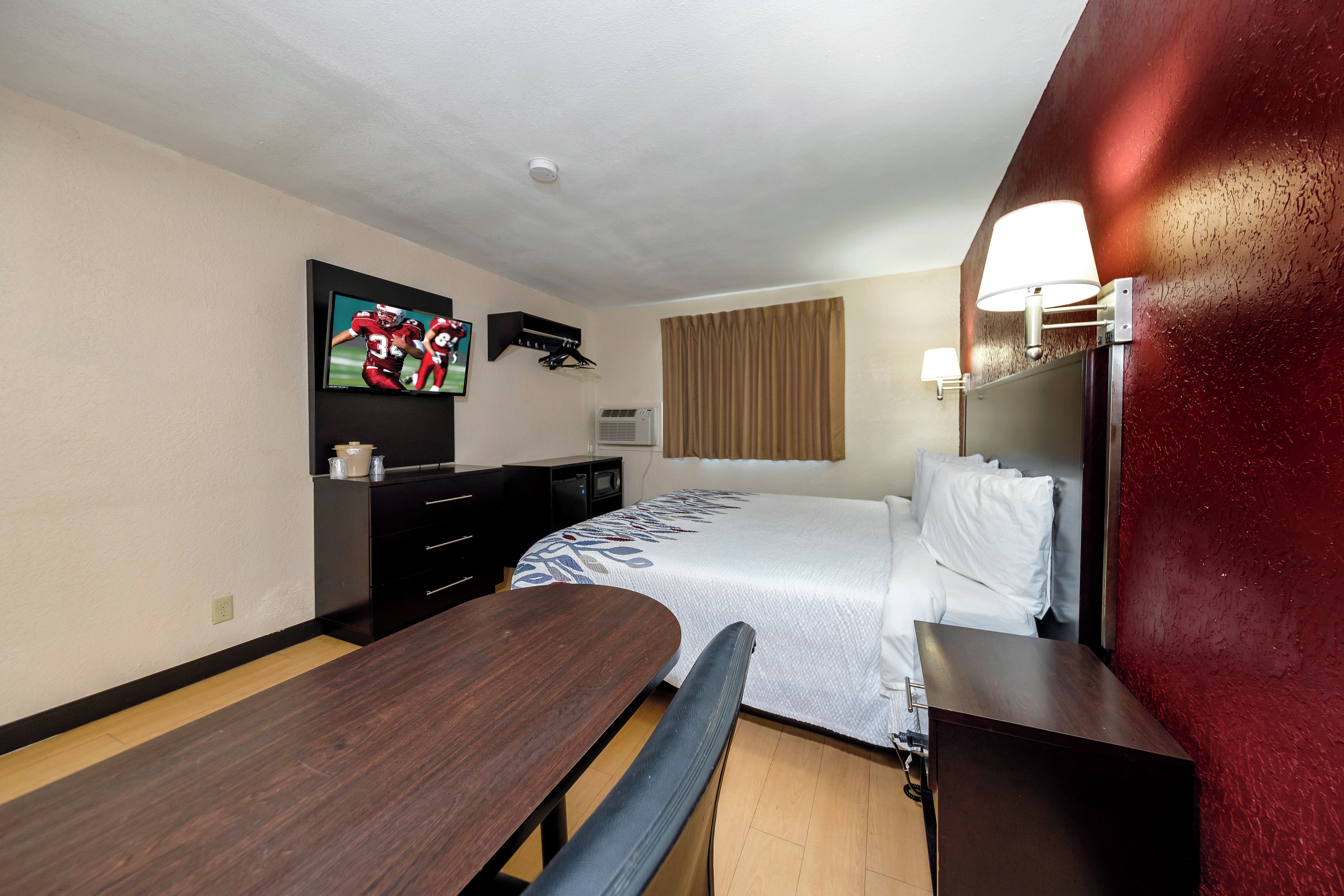 A hotel room with a bed, a desk, a chair, a TV mounted on the wall, and a window with curtains.