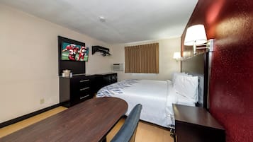 A hotel room with a bed, a desk, a chair, a TV mounted on the wall, and a window with curtains.