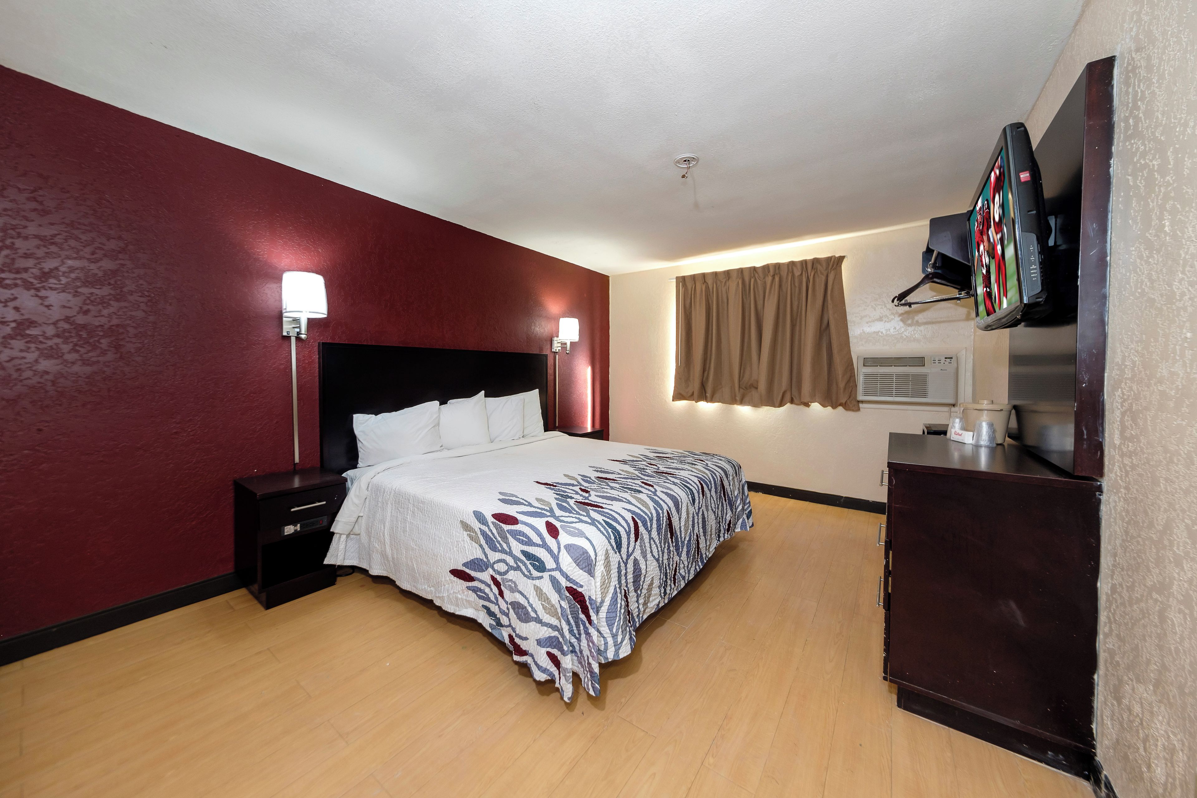 A hotel room with a bed, bedside lamps, a flat-screen TV mounted on the wall, a dresser, and a window with curtains.