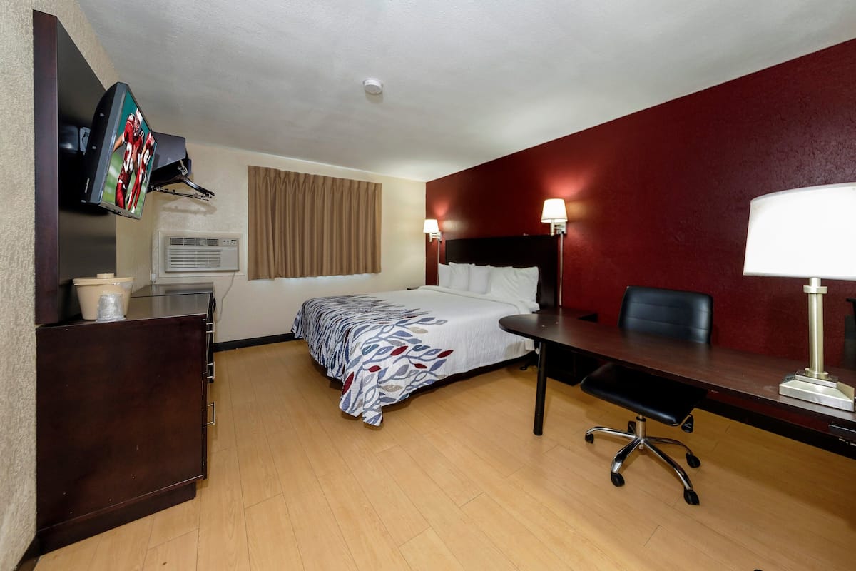 A hotel room with a bed, desk, chair, TV mounted on the wall, and a window with curtains.