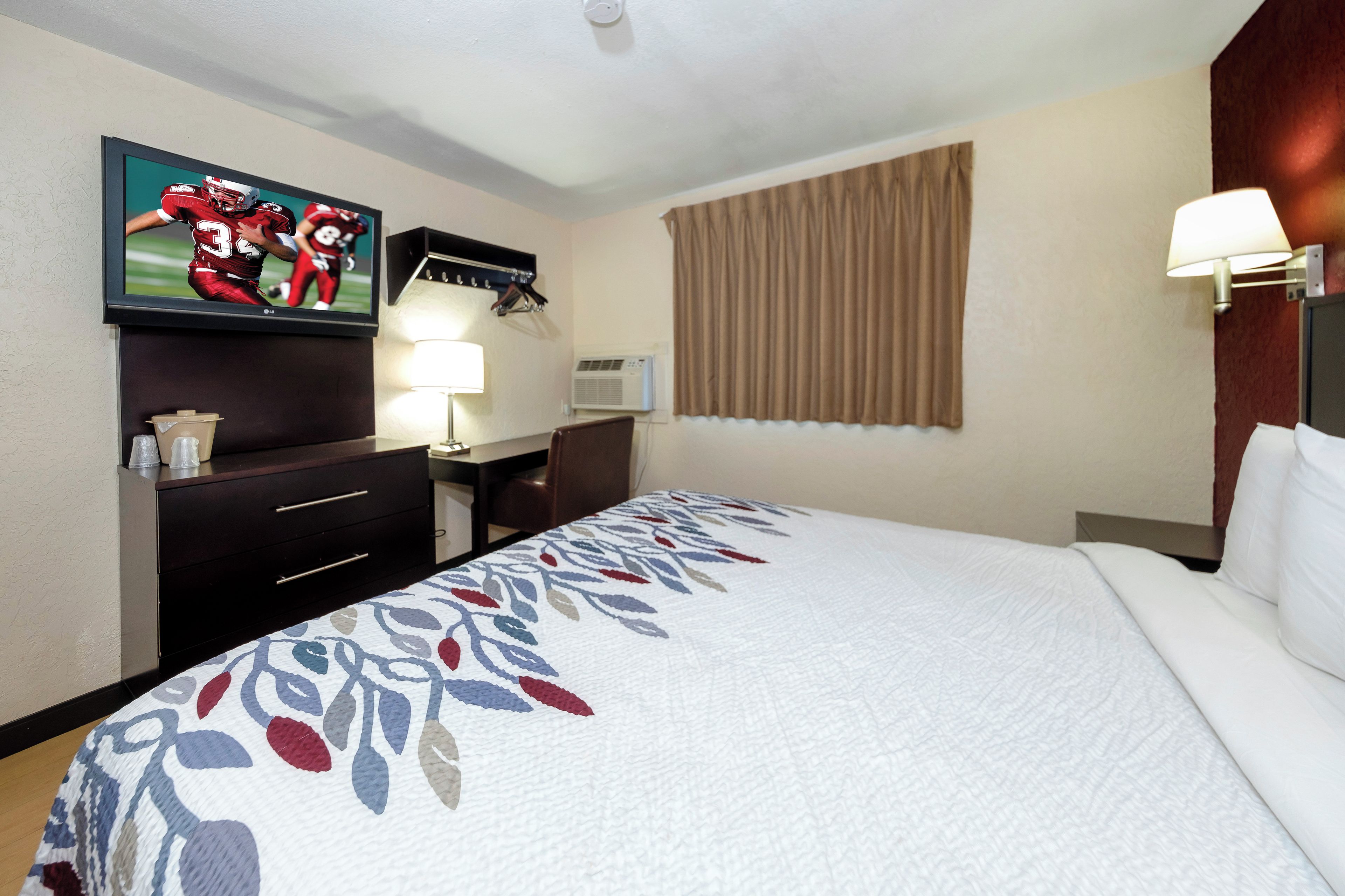 A hotel room with a bed, a TV mounted on the wall, a desk with a lamp, and a window with curtains.