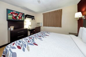 A hotel room with a bed, a TV mounted on the wall, a desk with a lamp, and a window with curtains. - Red Roof Inn Jacksonville - Cruise Port (Jacksonville)