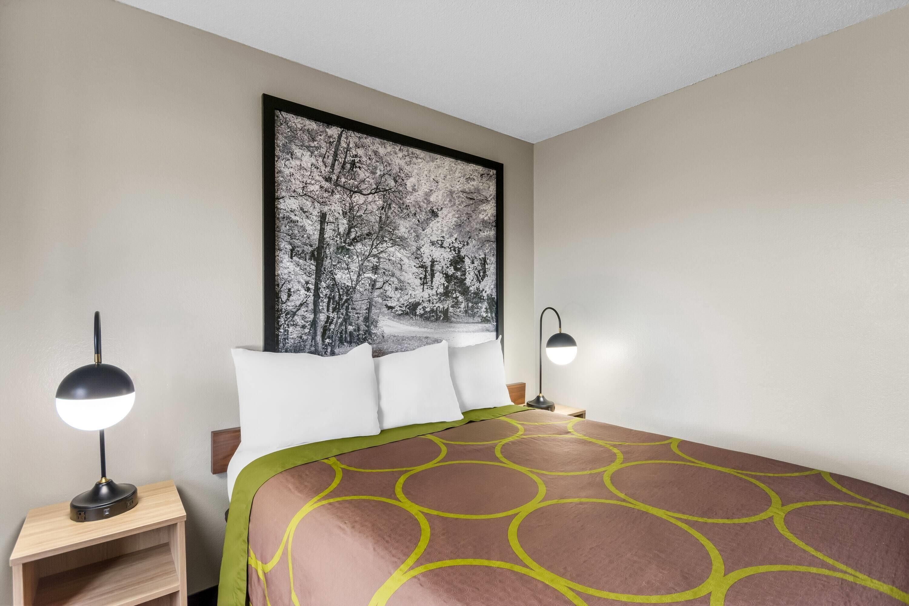 Habitación de hotel con cama, dos lámparas de noche, una mesita de noche y un cuadro grande en la pared.