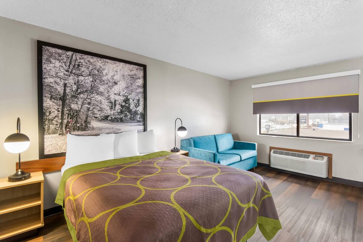 A hotel room with a bed, a blue armchair, a large window, and a wall-mounted air conditioner.