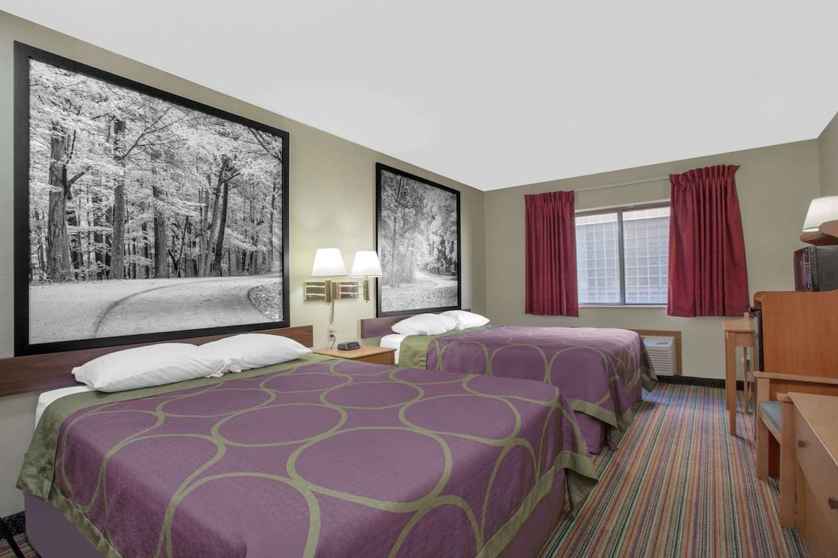 A hotel room with two beds, a desk, a chair, a window with red curtains, and two wall-mounted lamps.
