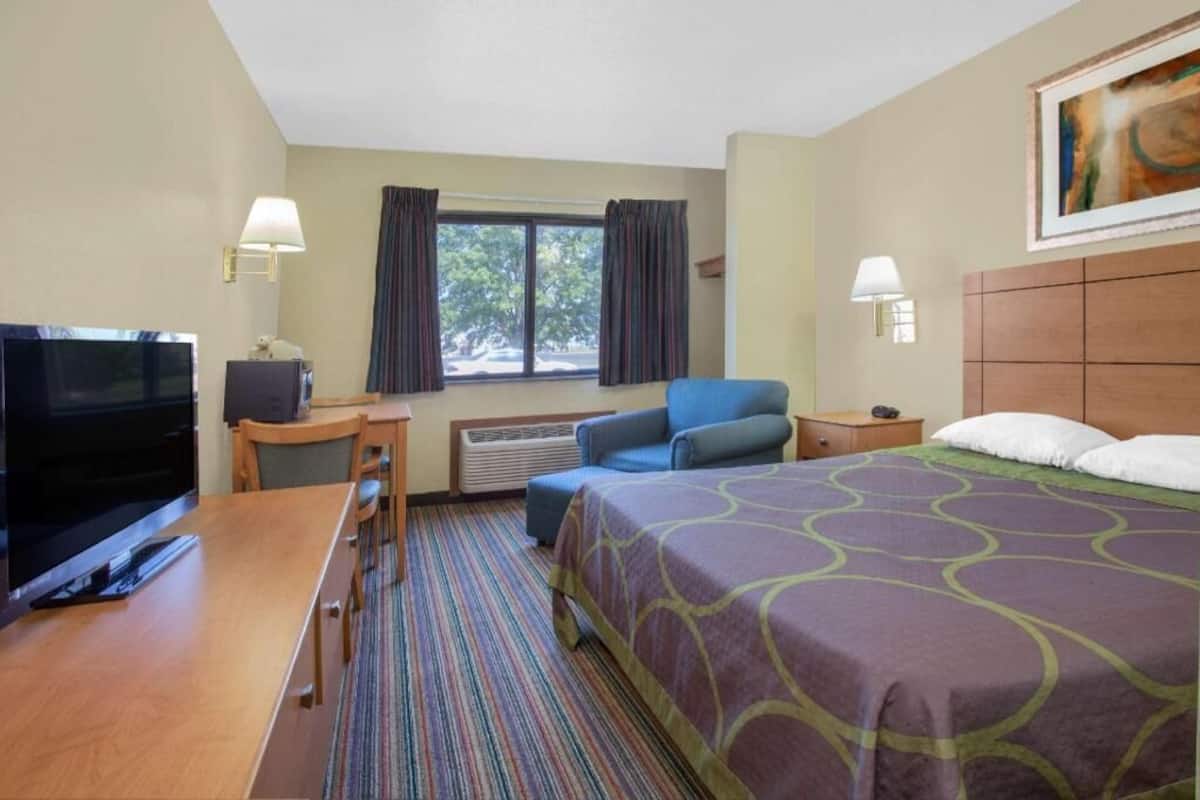 A hotel room with a bed, a desk with a TV, a chair, a window with curtains, and a painting on the wall.