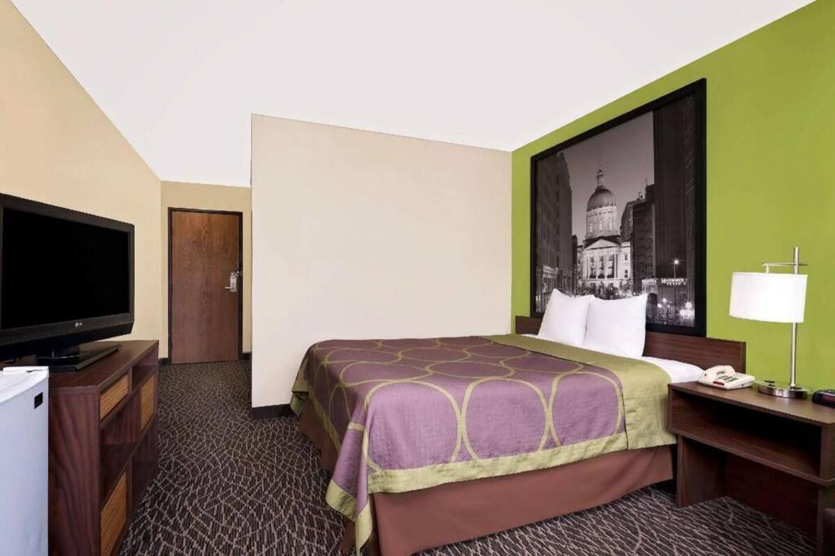 Un cuarto de hotel con una cama, televisión, escritorio con lámpara y un cartel de paisaje urbano en la pared.