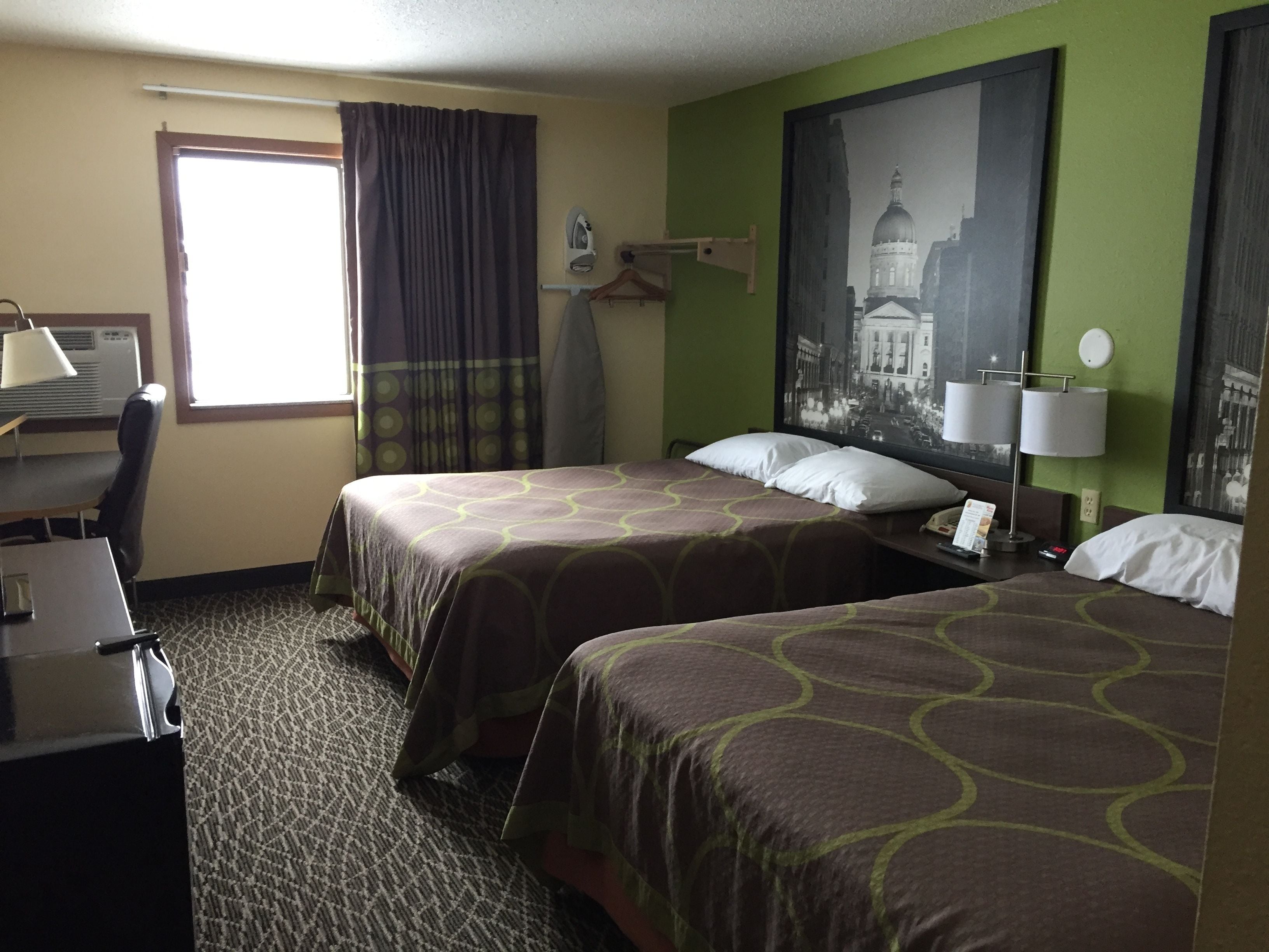 Quarto de hotel com duas camas, uma secretária, tábua de passar roupa, janela com cortinas e uma luminária na parede.