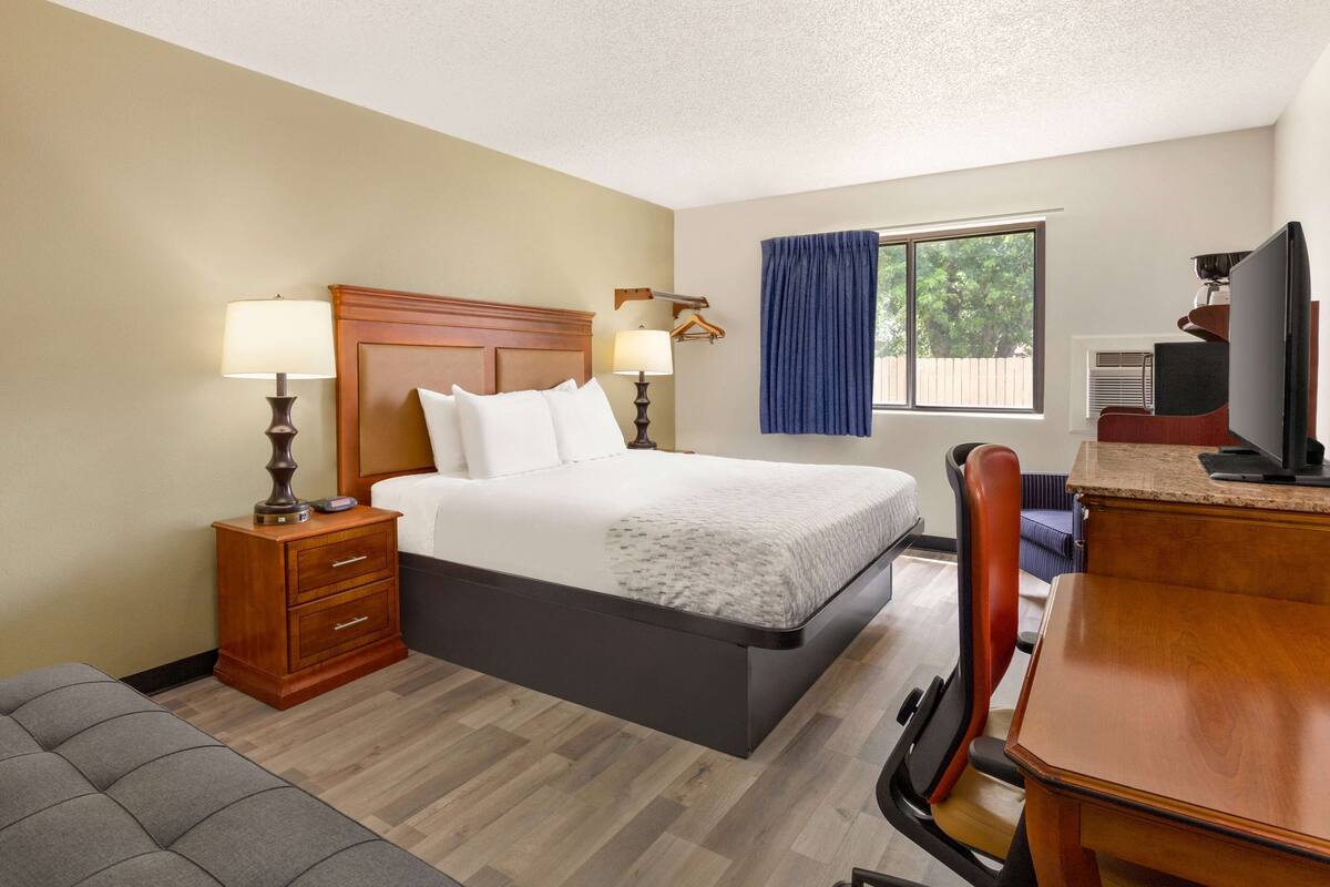 A hotel room with a bed, bedside tables, a desk, a chair, a TV, and a window with curtains.