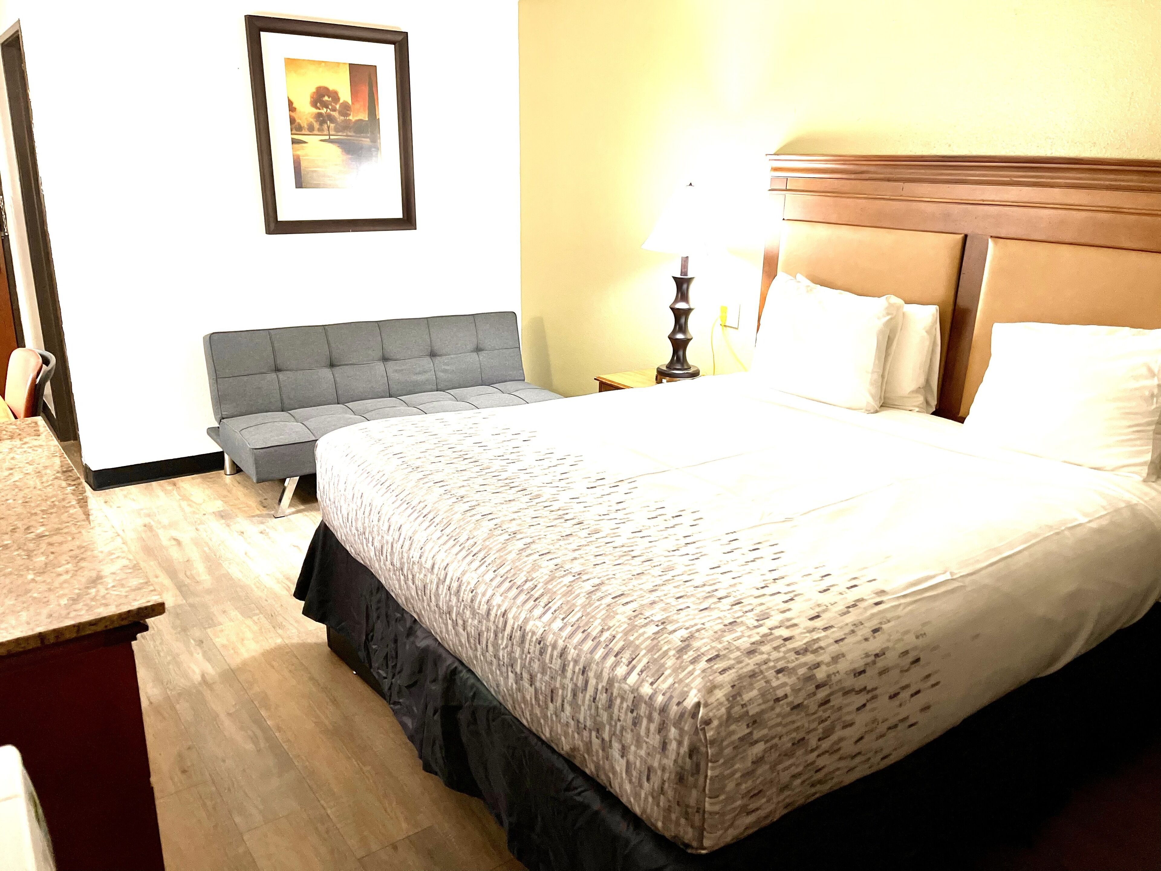 A hotel room with a large bed, a grey sofa, a small table, and a lamp.