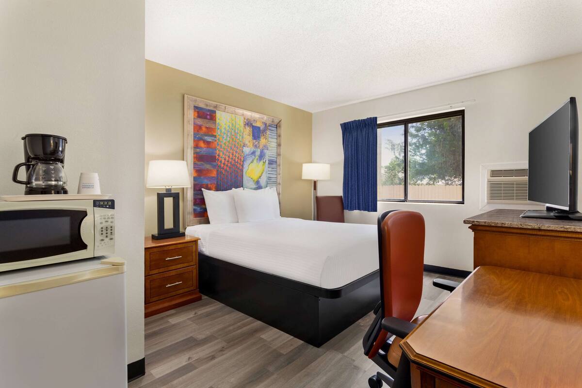 A hotel room with a bed, a desk, a television, a microwave, a coffee maker, and a window with curtains.
