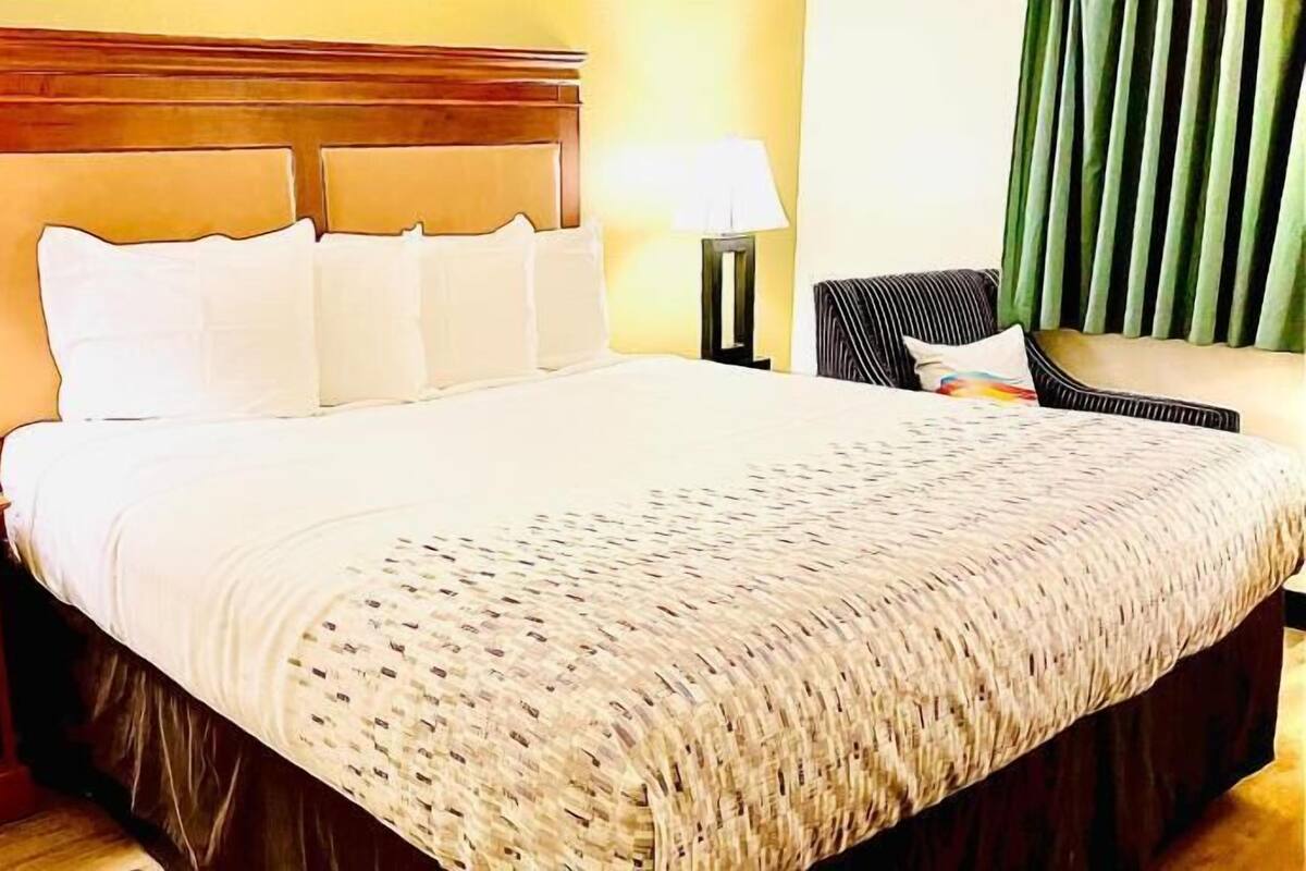 A hotel room with a large bed, wooden headboard, two bedside tables with lamps, a chair, and a window with blinds.