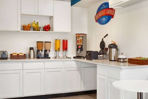 Cocina moderna con armarios blancos, cafetera, máquina para gofres y un cartel anunciando desayuno.