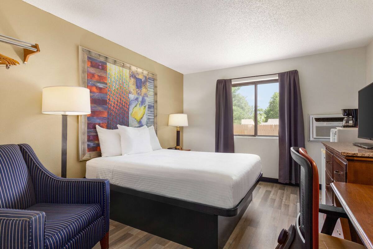 A hotel room with a bed, two bedside lamps, a desk with a computer, a chair, a TV, and a large abstract painting on the wall.
