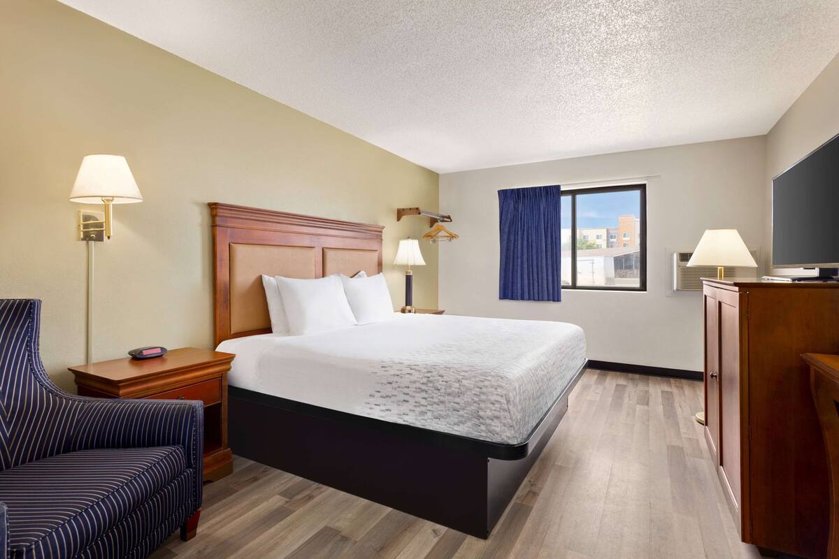 A hotel room with a bed, bedside tables, a chair, a TV, and a window with curtains.