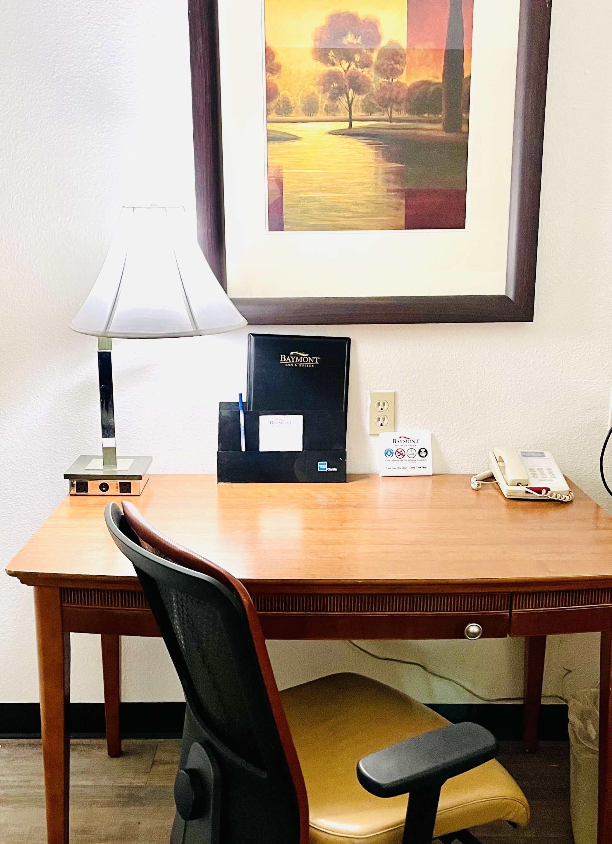 A hotel room desk with a lamp, a framed painting, a Baymont Inn Suites brochure, a phone, and a desk organizer.