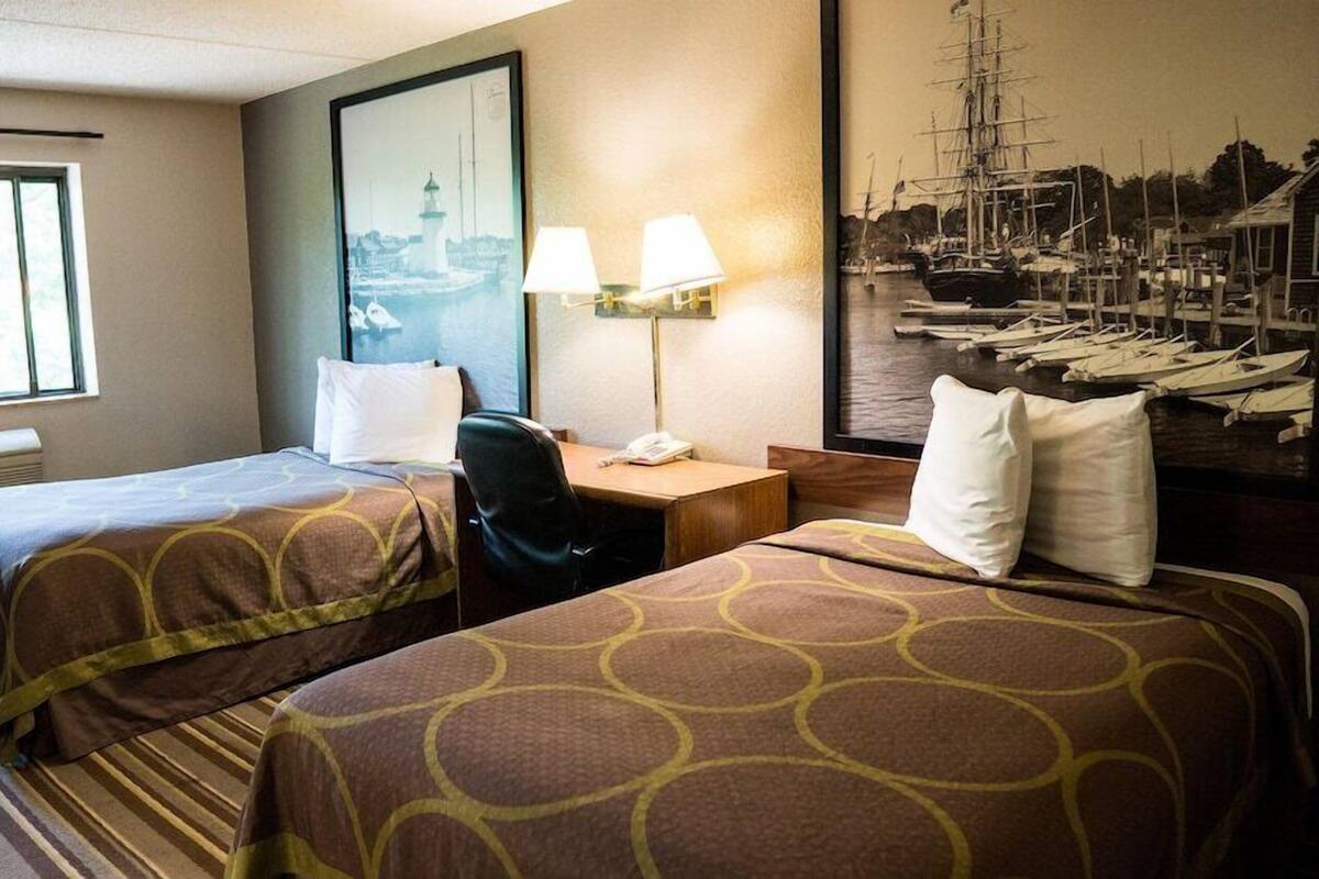 A hotel room with two beds, a desk, a chair, a lamp, a window, and a painting of a harbor scene.