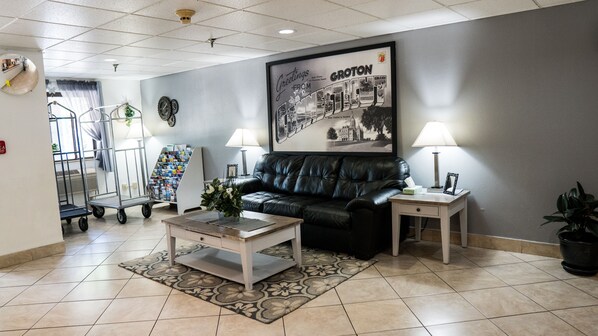 A lobby with a black leather sofa, a coffee table, a framed welcome poster, and a reception desk with a lamp. - Super 8 by Wyndham Groton (Groton)