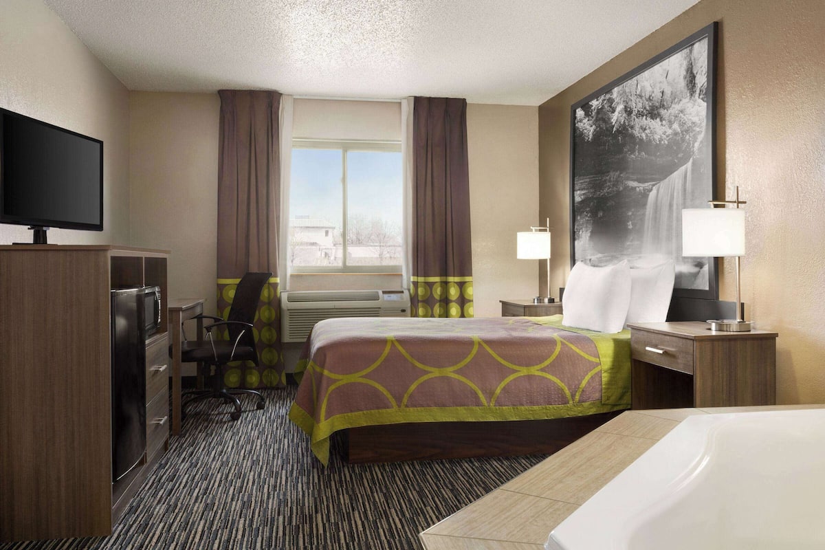 A hotel room with a bed, a desk with a computer, a TV, and a large window with curtains.