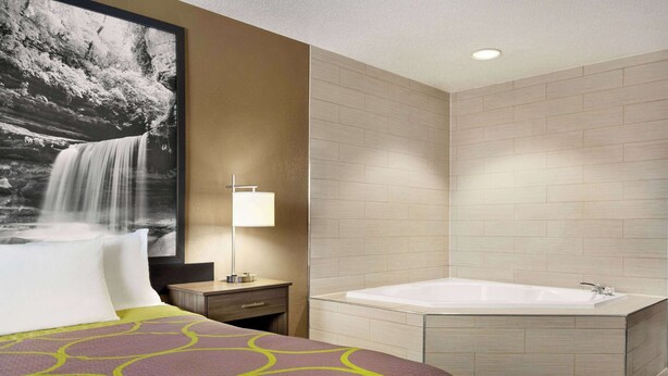 A hotel room with a large bed, a wall-mounted TV, a bedside table with a lamp, and a separate bathroom with a bathtub. - Super 8 by Wyndham Joliet I-55 N/Chicago (Joliet)