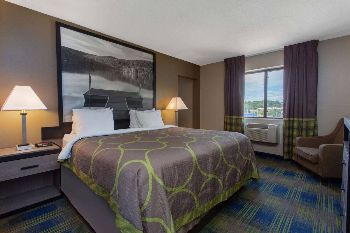 Une chambre d’hôtel avec un grand lit, deux tables de chevet, un bureau, une chaise, une fenêtre avec des rideaux et un tableau au mur.