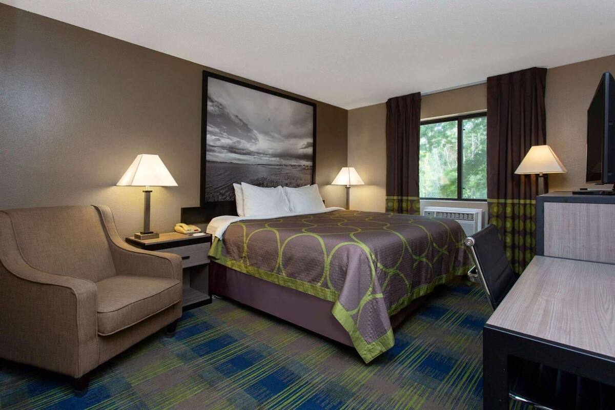 Une chambre d’hôtel comprenant un lit, deux tables de chevet, une chaise, un bureau et une fenêtre avec des rideaux.