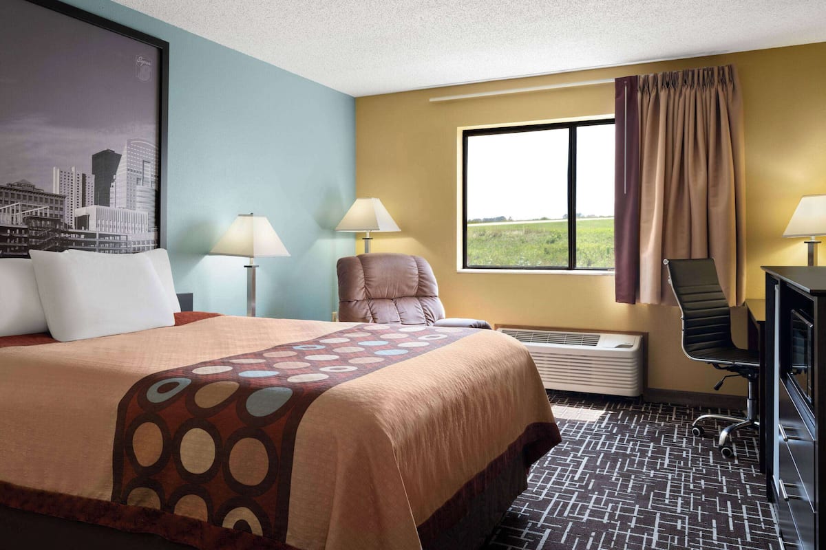 Une chambre d’hôtel avec un grand lit, deux fauteuils, un bureau, une fenêtre avec des rideaux et un tableau représentant un paysage urbain accroché au mur.